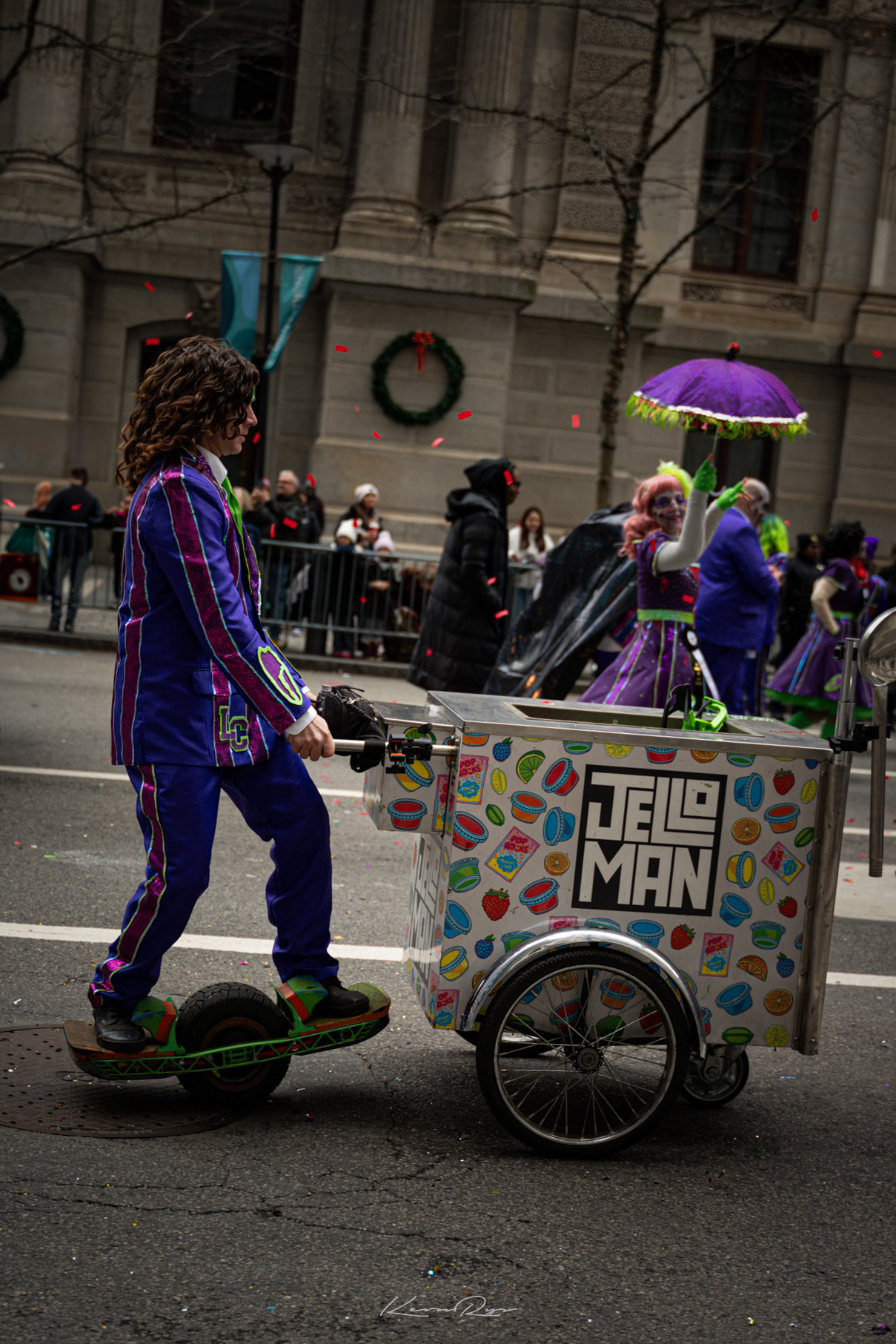 A Mummer in a striped purple suit rides a one-wheel board while pushing a Jello Man cart past confetti and parade watchers