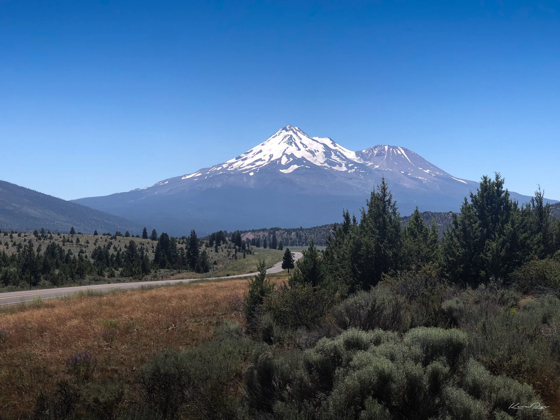 Snow-capped Mt. Shasta under a clear blue sky, with pine trees, sagebrush plus a two-lane road in the foreground