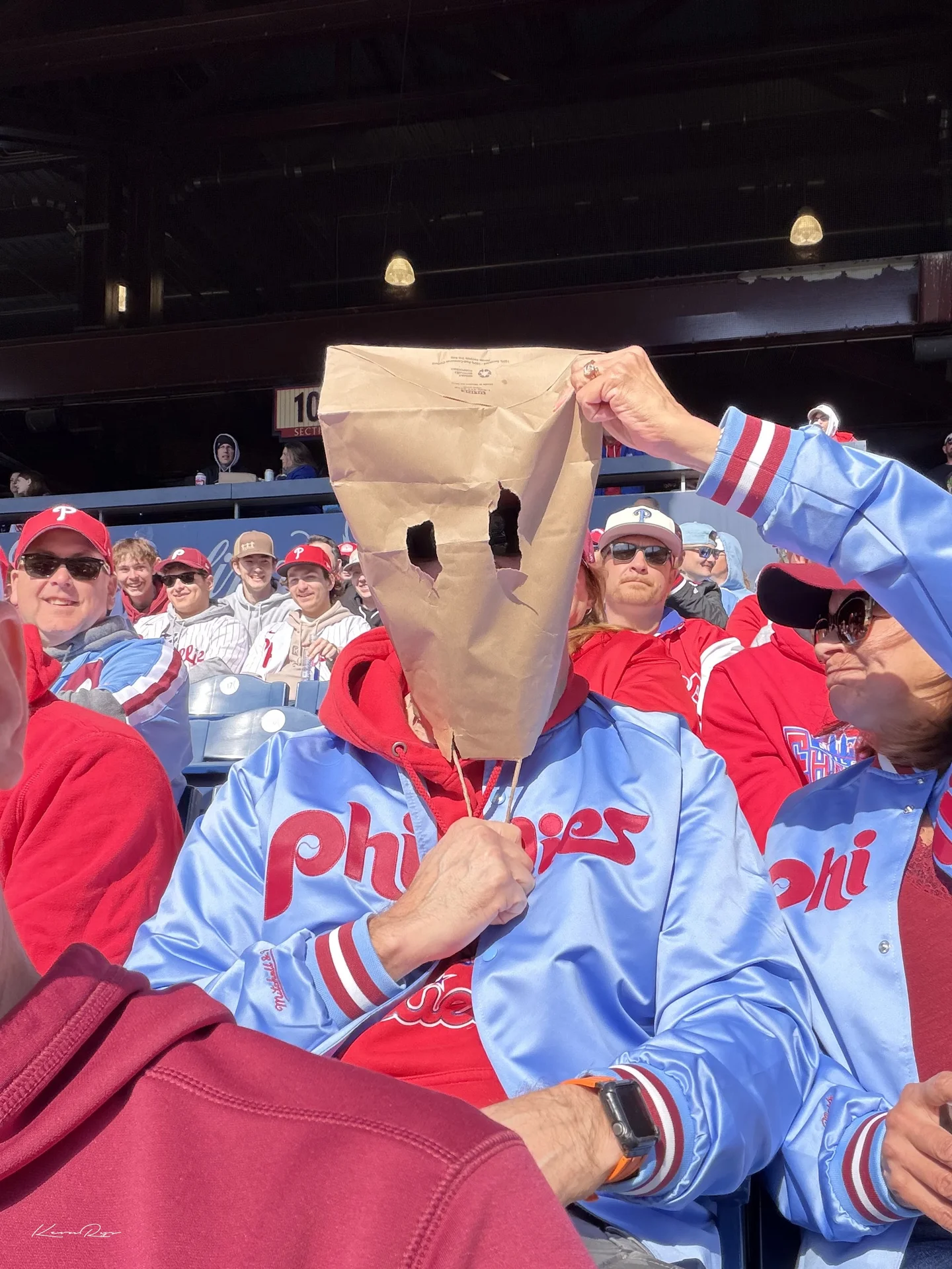 A fan in a powder blue jacket wears a brown paper bag with eye holes as a neighbor tugs at its top, fans grinning behind